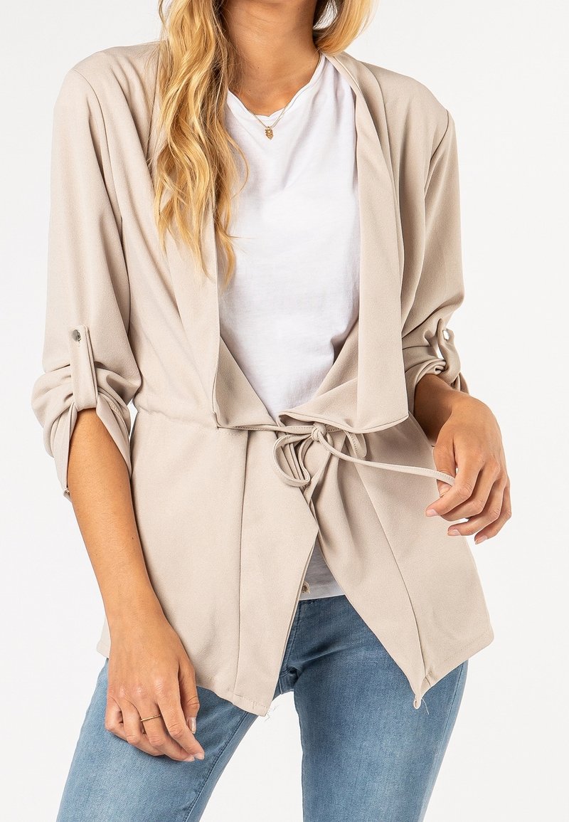 Femme portant une veste beige ouverte devant avec ceinture à nouer, t-shirt blanc, jean bleu et un petit collier avec pendentif.