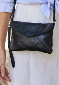 Bolso de piel negra tipo bandolera con una solapa triangular y una textura sutil. Presenta una correa larga y una forma rectangular delgada.