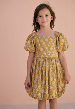 Junges Mädchen mit dunklem Haar, das ein senfgelbes Kleid mit Puffärmeln und weißem Blumenmuster trägt, vor einem schlichten pfirsichfarbenen Hintergrund stehend.
