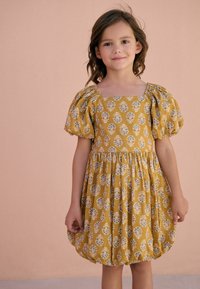Mosterdkleurige jurk met pofmouwen, vierkante hals en geplooide taille. Heeft een witte bloemenprint en een afgeronde zoom.