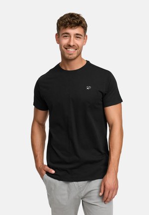 Young man smiling, wearing a black short-sleeve t-shirt with small white logo on chest, light gray pants, hand in pocket, standing against white background.
