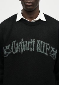 Μαύρο πουλόβερ με ξεθωριασμένο γοτθικό κείμενο "Osthavsti Cup" και δύο έμβληματικά λιοντάρια, φορεμένο πάνω από ένα λευκό πουκάμισο με γιακά.