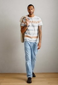 Mann som går innendørs iført en stripete T-skjorte, lyseblå jeans, mørke sko, med en lys brunt jakke over skuldrene.