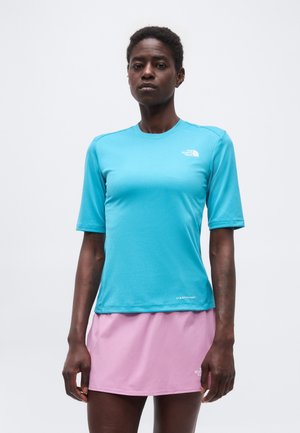 Személy türkiz színű, rövid ujjú sportpólót és világos rózsaszín szoknyát visel, egyszínű fehér háttér előtt állva.