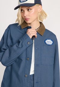 Blaue Jeansjacke mit einem braunen Cordkragen, ausgestattet mit einer Brusttasche, Druckknöpfen und einem gestickten Logopatch.