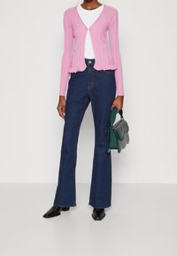 Roze geribbelde cardigan met een peplumzoom, gecombineerd met donkerblauwe uitlopende jeans. Het model houdt een groene handtas vast. Zwarte pumps met een spits toelopende neus maken de look compleet.