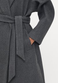 Cappotto grigio in misto lana con vita accentuata da una cintura, maniche lunghe e tasche laterali. Il tessuto ha un aspetto morbido e testurizzato.