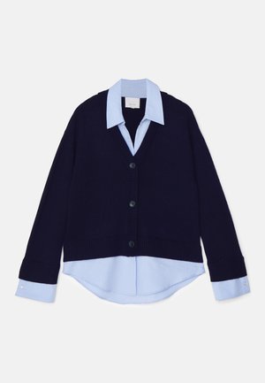 Marine blauwe gebreide cardigan met een V-hals en geribbelde randen, met daaronder een lichtblauw collared shirt. Vier zwarte knopen sieren de voorkant.