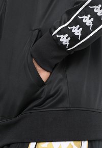 Black athletic jacket featuring a textured fabric, side pocket, and white logo accents along the sleeve. Contrast ribbed cuffs are visible.