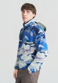 Νέος άνδρας που φοράει μπλε-λευκό φλις μπουφάν με πατιλέτα με μισά κουμπιά και πράσινο σακίδιο στην πλάτη του.