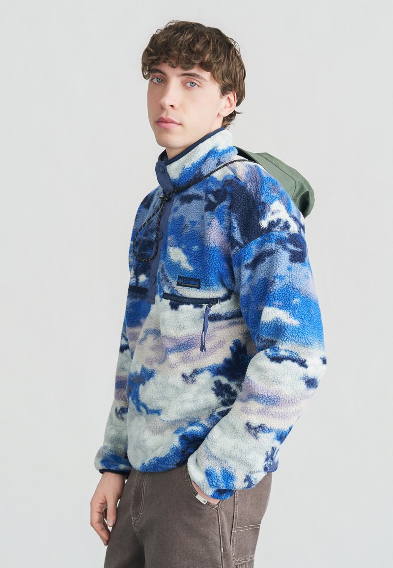 Νέος άνδρας που φοράει μπλε-λευκό φλις μπουφάν με πατιλέτα με μισά κουμπιά και πράσινο σακίδιο στην πλάτη του.