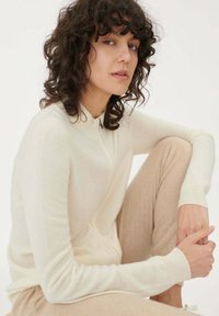 Krämfärgad zip-up tröja med hög krage, ribbade manschetter och mjuk textur, kombinerad med ljusbeige byxor. Närbild på en person.