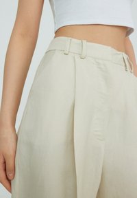 Licht beige geplooide broek met een hoge taille, voorzien van een platte voorkant, zijzakken en een zachte linnen textuur. Inclusief verstelbare riemlussen.