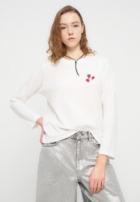 Λευκό μακρυμάνικο T-shirt με μαύρο ντεκολτέ και τρία κόκκινα γραφικά με φράουλες, σε συνδυασμό με γυαλιστερό ασημένιο παντελόνι με φαρδιά πόδια.