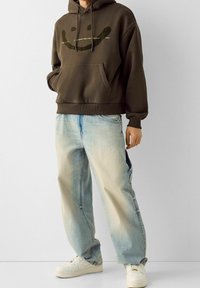 Felpa con cappuccio marrone con grafica sorridente e testo, abbinata a jeans larghi azzurri. Modello con scarpe da ginnastica bianche, in piedi su uno sfondo neutro.