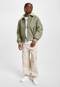 Ung mand med fletninger, iført en olivengrøn jakke med pelskrave, hvid t-shirt, beige cargo-bukser og hvide sneakers, stående.
