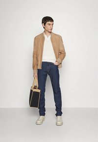 Tan semsket bomberjakke over en hvit strikkegenser, kombinert med mørkeblå jeans og beige joggesko. Han holder en handleveske med svart front.