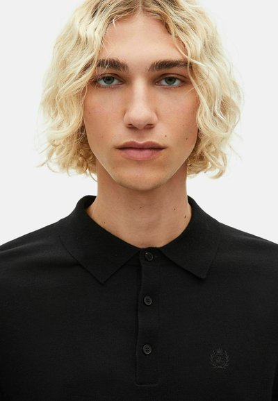 Polo nero con colletto classico e quattro bottoni, impreziosito da un logo discreto sul lato sinistro del petto. Tessuto liscio e texture morbida.