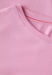 Světle růžová bavlněná látka má kulatý výstřih s červeným švem podél okraje, který ukazuje hladkou texturu a jednoduchý design.