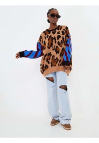 Maglione oversize a stampa leopardata marrone con maniche a motivo blu e rosso, abbinato a jeans strappati blu chiaro e scarpe marroni.