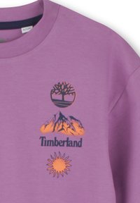 Paarse sweatshirt met zwarte en oranje grafische ontwerpen, waaronder een boom, bergen, de tekst "Timberland" en een zon. Zachte textuur.