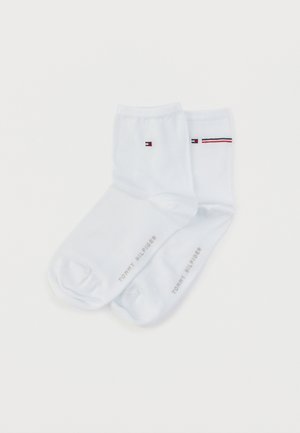 Chaussettes blanches en coton coupées à la cheville, avec un petit logo rouge, blanc et bleu, une bande rayée contrastante et le nom de la marque imprimé sur le côté.