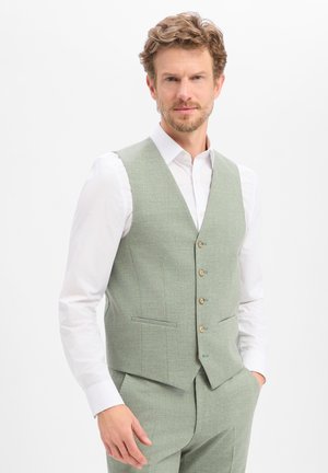 Mann mit lockigem Haar, der eine hellgrüne Anzugweste und passende Hose über einem weißen Hemd trägt, steht mit einer Hand in der Tasche.