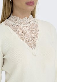 White sweater featuring a high lace collar with intricate floral patterns, ribbed texture, and a V-shaped neckline.