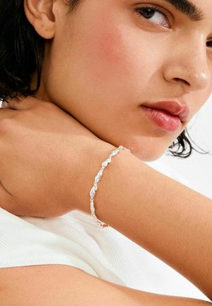 Srebrna narukvica s valovitim dizajnom i teksturiranim detaljima, nošena na zapešću, s neutralnom pozadinom i mekanim, prirodnim osvjetljenjem.