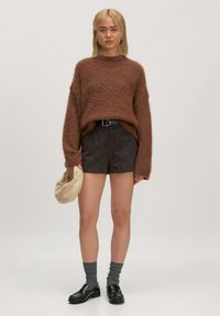 Vrouw die staat en een bruine harige trui draagt, gestructureerde donkergekleurde shorts met een riem, grijze sokken, zwarte loafers, terwijl ze een beige zachte clutch tas vasthoudt.