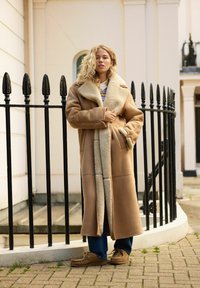 Kurtka shearling w kolorze tan z dużym kołnierzem i przednimi kieszeniami, w połączeniu z niebieskimi dżinsami i jasnobrązowymi butami. Teksturowany materiał i luźny krój.