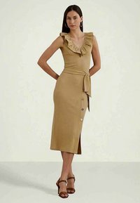 Žena stojí s rukami za chrbtom, oblečená v pieskovohnedom bezrukávovom midi šatách s volánovým výstrihom, bočnými gombíkmi, opaskom v páse a hnedými vysokými sandálmi.