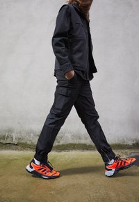 Černá bunda s fleecovou podšívkou a zipovými kapsami, černé kalhoty typu cargo a jasně oranžové sportovní boty s černými a bílými detaily.