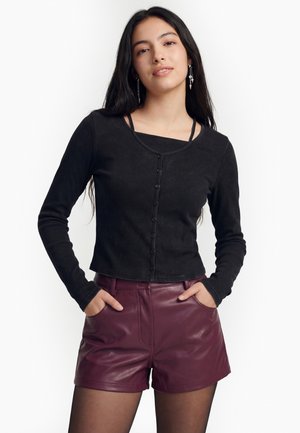 Ung kvinde med langt mørkt hår iført sort langærmet knapbluse og bordeaux lædershorts, hænderne i lommerne, foran hvid baggrund.