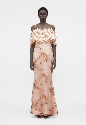 Γυναίκα που φοράει ανοιχτό ροζ φόρεμα έξω από τους ώμους με 3D φλοράλ εφαρμογές και κεντημένα κλαδιά, ποζάροντας σε απλό λευκό φόντο.