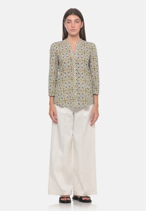 Donna che indossa una camicetta a maniche lunghe con fantasia, pantaloni bianchi a gamba larga e sandali neri, in piedi davanti a uno sfondo uniforme.