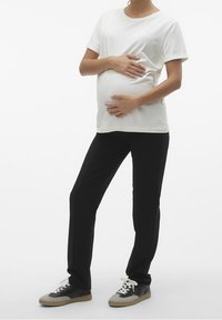 Vit kortärmad skjorta och svarta byxor på en gravid figur, händerna vilande på magen, med gråa och svarta sneakers. Enkelt design och passform.