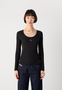 Zwarte lange mouwen top met een geribbelde textuur en een ronde halslijn, voorzien van een klein logo-opdruk in het midden. Gecombineerd met donkere denimjeans.