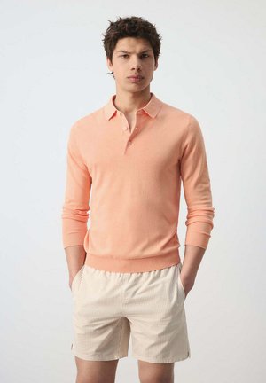 Joven con una camiseta polo de manga larga color melocotón y pantalones cortos a rayas beige claro, de pie con las manos en los bolsillos sobre un fondo liso.