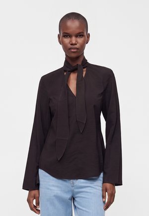 Femme aux cheveux courts porte un chemisier noir à manches longues avec un foulard noué au cou et un jean bleu clair sur un fond uni.