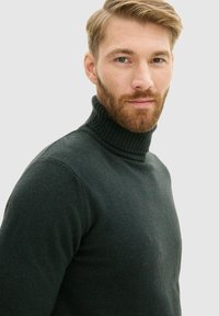 Dunkelgrüner Rollkragenpullover aus weichem Strickmaterial, mit einem gerippten Kragen und taillierter Passform sowie subtilen Texturdetaillierungen.