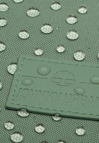 Zöld textúrájú szövet, amelyen tiszta vízcseppek láthatók, és egy téglalap alakú címke, amelyen a "JOHNNY URBAN" felirat található, szintén zöld színben, beleolvadva a háttérbe.