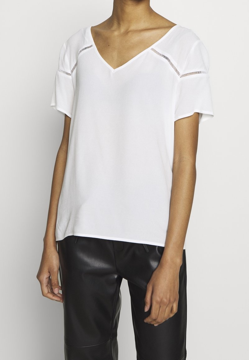 Personne portant un chemisier blanc à col en V avec manches courtes et un pantalon en cuir noir, debout devant un fond clair uni.