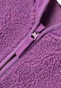 Veste en polaire violette avec une surface texturée et duveteuse. Elle présente une fermeture éclair centrale avec un bord en tissu lisse et une couche intérieure contrastante.