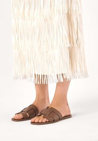 Hnedé kožené šľapky s geometrickým dizajnom pásku. Nosí sa s bielou sukňou s strapcami, pričom ukazuje plochý, otvorený štýl.