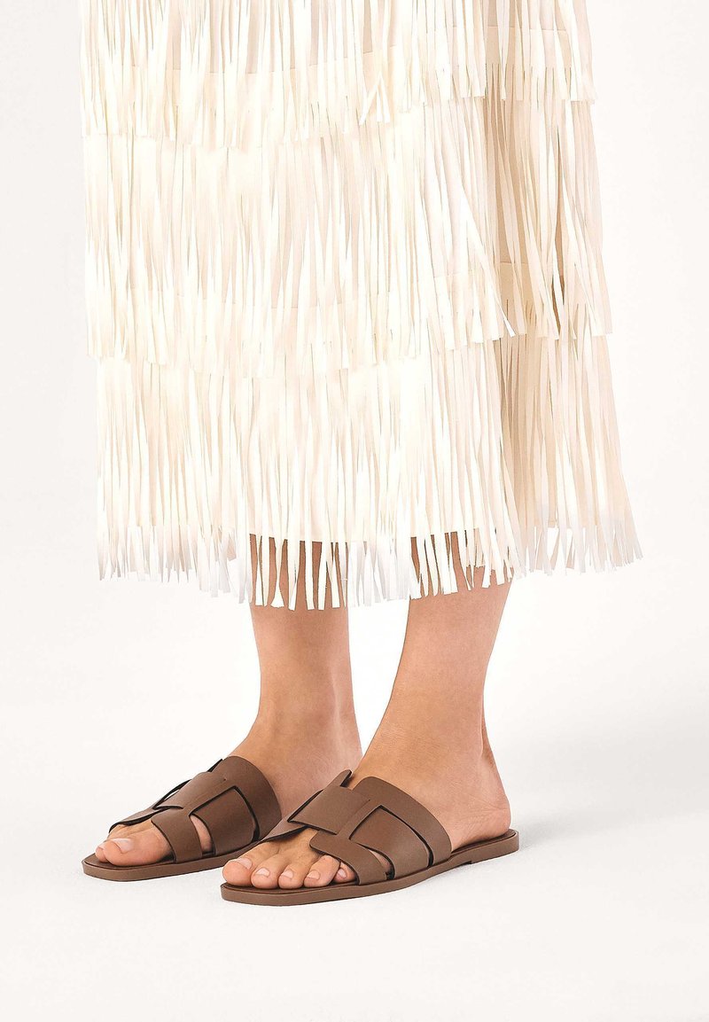 Hnedé kožené šľapky s geometrickým dizajnom pásku. Nosí sa s bielou sukňou s strapcami, pričom ukazuje plochý, otvorený štýl.