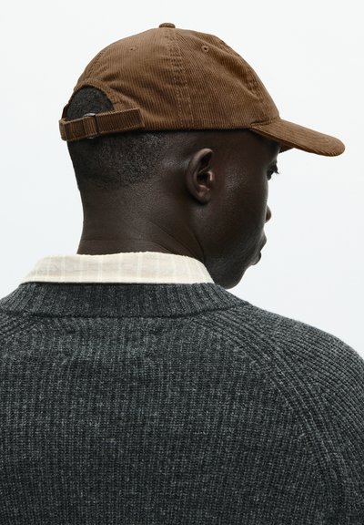 Barna corduroy sapka ívelt peremmel és állítható pánttal a hátsó részen, sötétszürke kötött pulóverrel és világos színű galléros inggel viselve.