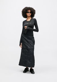 Čierny top s dlhým rukávom s bielym prešívaním, v kombinácii s čiernou, členkovou prešívanou sukňou. Modelka má na sebe tmavé slnečné okuliare a čierne topánky.