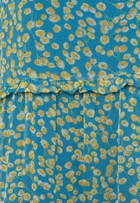 Tissu bleu ornée d'un motif floral en jaune et blanc, avec un ourlet froncé. La texture semble douce et légèrement côtelée.