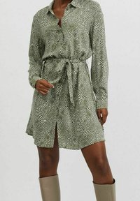 Kvinna i en olivgrön skjortklänning med vitt prickmönster, knytning vid midjan, långa ärmar och beige knähöga stövlar.